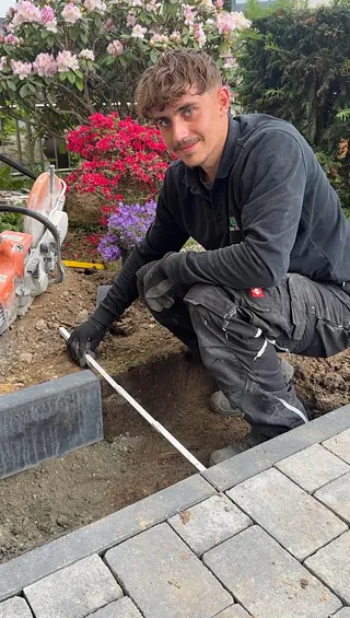 Eine Person pflanzt bunte Blumen in ein Beet mit Steinen, umgeben von grünen Sträuchern und Bäumen.